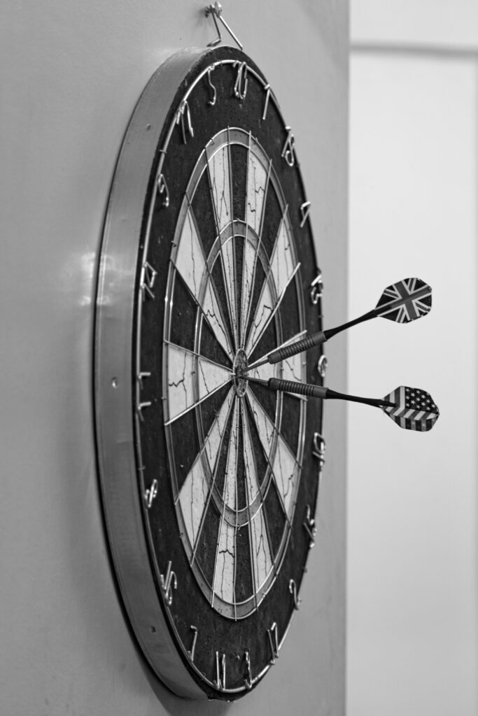 A monochrome side view of a dartboard with three darts, including UK and US flags.